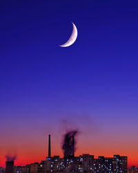 Crescent moon over smoking chimneys on apartment buildings at dusk