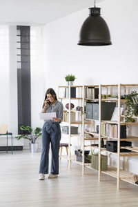 Smiling businesswoman talking on mobile phone while looking at plan standing against rack in office