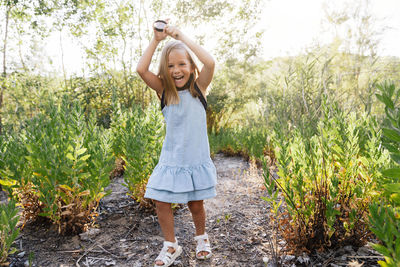 Portrait of happy girl holding magnifying glass standing on field amidst plants in forest