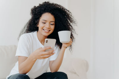 Young woman using mobile phone while drinking coffee at home