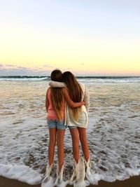 Rear view of friends with arms around standing at beach against sky during sunset