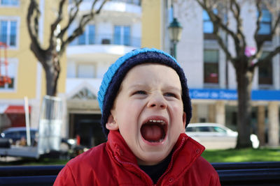 Emotional boy with his mouth wide open. little boy screaming in a red jacket and blue hat on street