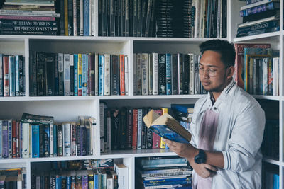 Young man reading book against shelves