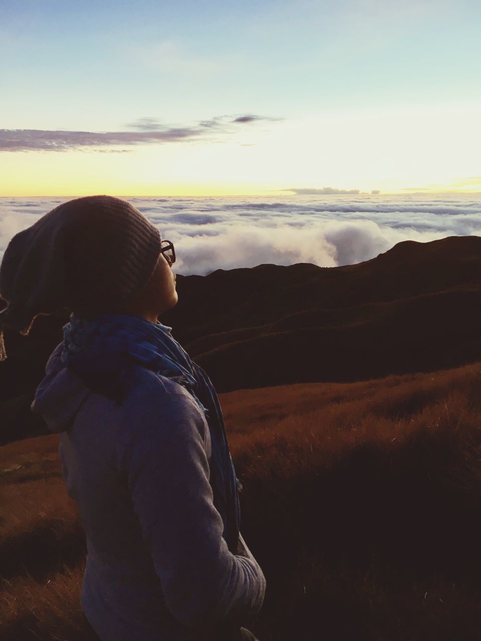 REAR VIEW OF WOMAN STANDING ON LANDSCAPE AGAINST SKY AT SUNSET