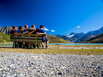 People sitting on bench by mountain against blue sky