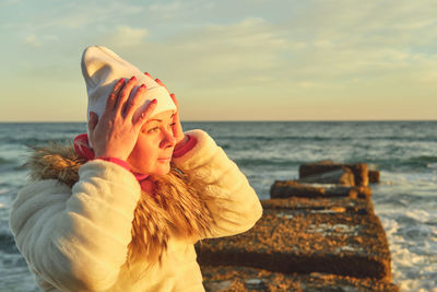 A woman with blond hair in a hat and a fur coat walks along the sea along a stone breakwater