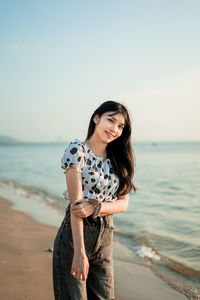 Portrait of smiling young woman standing at beach against clear sky