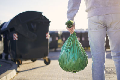 Man walking with rubbish. rear view of person carrying plastic bag against garbage cans on street.