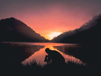 Silhouette man crouching against lake during sunset