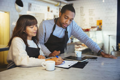 Female employee discussing over clipboard with male coworker at table in cafe