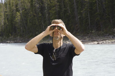 Mature man looking through binoculars while standing against lake in forest