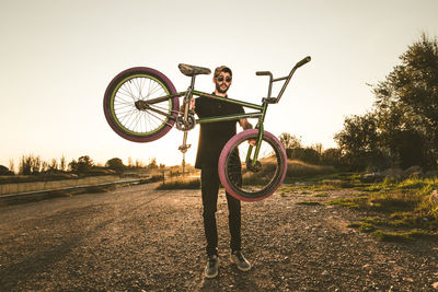 Portrait of young man carrying bicycle standing on field against clear sky during sunset