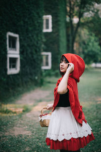 Woman in little red riding hood costume holding basket while standing against building