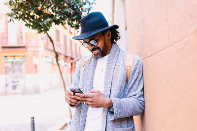 Hispanic male in stylish clothes with hat and sunglasses leaning on wall and browsing cellphone while standing on pavement on city street