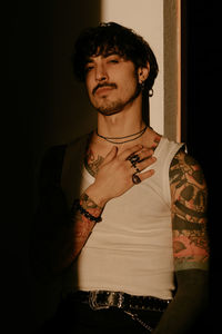 Young stylish guy with mustache and tattoos wearing white tank top and leaning against wall with back while looking at camera