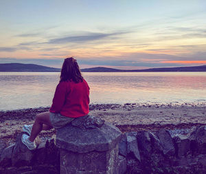 Rear view of woman sitting on rock by sea against sky
