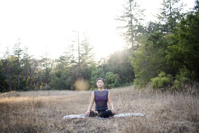 Woman meditating while sitting on exercise mat against clear sky at field