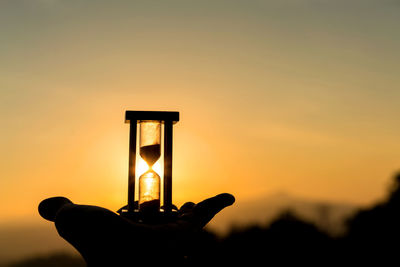 Silhouette hand holding hourglass against sky during sunset