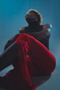 Confident african american female model in stylish outfit sitting on floor in dark studio against blue background and looking away
