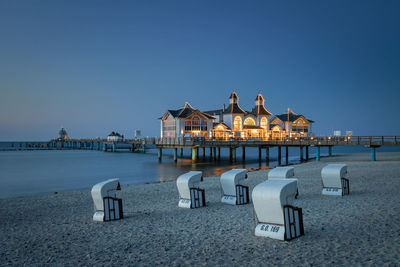 Hooded chairs at beach by stilt houses over sea against clear sky