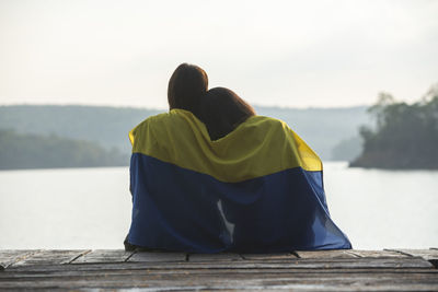 Rear view of girls wearing ukrainian flag sitting on pier by lake