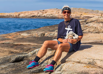 Full length portrait of senior woman with toys sitting on rock at beach during sunny day