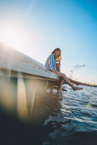 Side view of young woman sitting on pier in lake against clear sky during sunny day