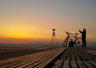 Silhouette man photographing bicycle against sky during sunset