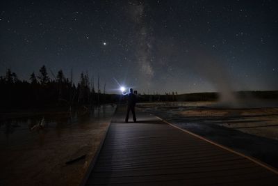 Rear view of man standing on illuminated street against sky at night