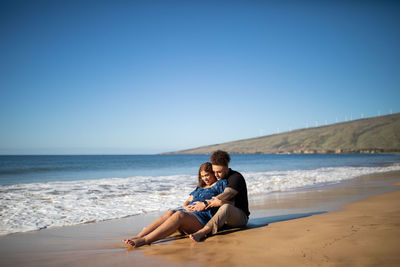 Side view of a man and woman sitting at beach against clear sky