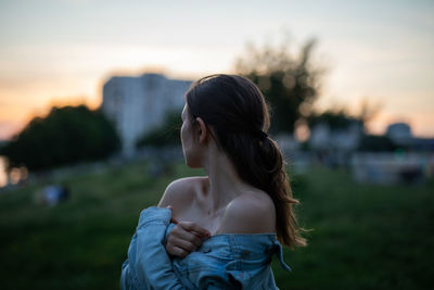 Woman wearing denim jacket standing against sky during sunset