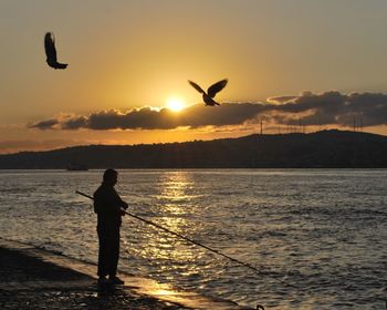 Silhouette man standing in sea against sky during sunset