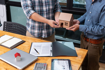 Midsection of real estate agent and customer holding model house at office