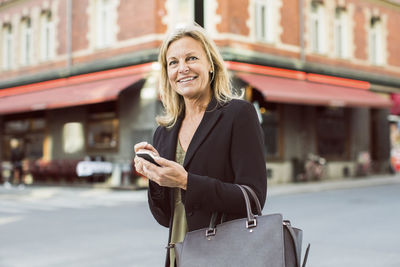 Happy businesswoman looking away while using mobile phone on city street