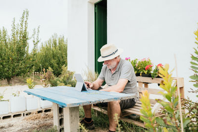 Elderly man in casual clothes and hat sitting on wooden bench at shabby table and browsing data on tablet during break in garden outside house on summer day in countryside