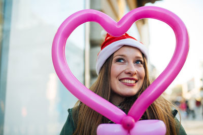 Smiling young woman wearing santa hat by heart shape balloon