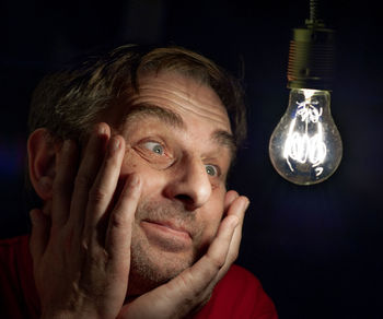 Close-up of mature man looking at illuminated light bulb against black background