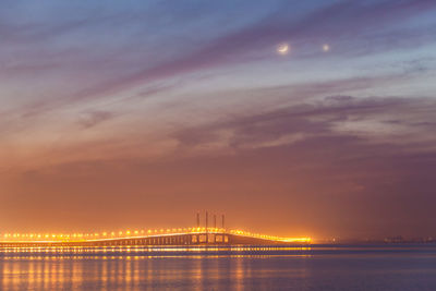 Illuminated bridge over sea against cloudy sky at night