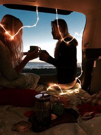 Female friends having drink while sitting in vehicle at beach during sunset