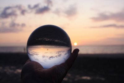 Close-up of hand holding crystal ball against sea during sunset