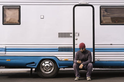 Full body of young bearded male traveler in casual clothes and sunglasses sitting on step of camper van and messaging on smartphone