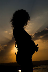 Silhouette pregnant woman standing at beach against sky during sunset