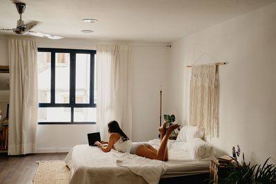 Cheerful millennial female in nightwear lying on bed and surfing internet on laptop while spending weekend morning in cozy bedroom with minimalist interior