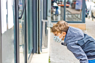 Kid wearing protective face mask and peeking through store window during coronavirus pandemic.