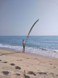 Shirtless boy holding dry palm leaves at beach against clear sky
