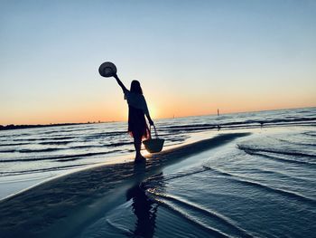 Full length of silhouette woman with hat standing at beach against sky during sunset