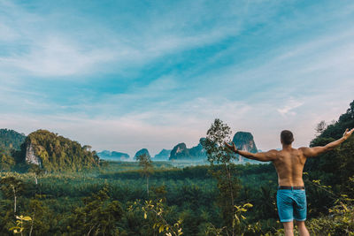 Rear view of shirtless young man with arms outstretched standing in forest against sky