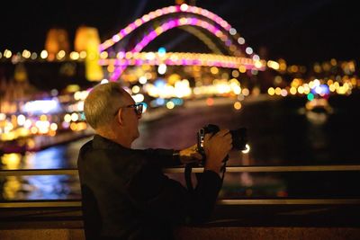 Side view of man sitting by illuminated railing at night