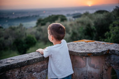 Side view of boy looking away while sitting on retaining wall against sky during sunset