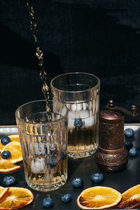 Pouring lemonade into glasses decorated with blueberries and dry orange on black background